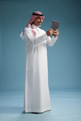 A portrait of a Saudi Arabian Gulf man wearing a white thobe and a shemagh using a digital tablet, full body, with a blue background.