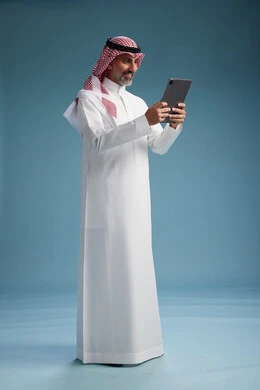 بورتريه لرجل عربي سعودي  خليجي يرتدي الثوب  الابيض والشماغ يستخدم جهازاً لوحياً رقمياً, كامل الجسم, خلفيه زرقاء