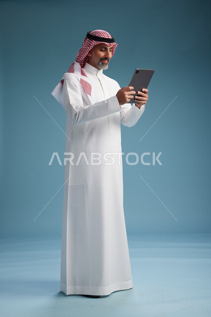 بورتريه لرجل عربي سعودي  خليجي يرتدي الثوب  الابيض والشماغ يستخدم جهازاً لوحياً رقمياً, كامل الجسم, خلفيه زرقاء