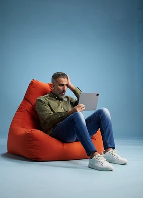 A portrait of a Saudi Arabian Gulf man wearing casual attire, focused while using a tablet while sitting on a chair among orange foliage.