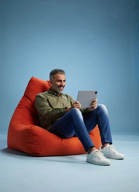 A portrait of a Saudi Arabian Gulf man wearing casual attire, focused while using a tablet while sitting on a chair among orange foliage.