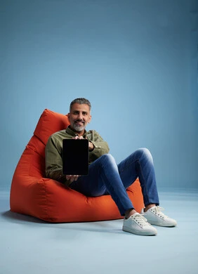A portrait of a Saudi Arabian Gulf man wearing casual attire, focused while using a tablet while sitting on a chair among orange foliage.