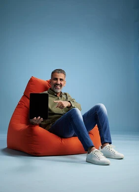 A portrait of a Saudi Arabian Gulf man wearing casual attire, focused while using a tablet while sitting on a chair among orange foliage.