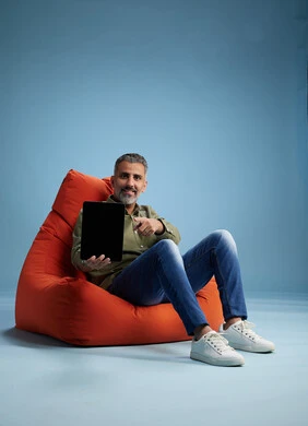A portrait of a Saudi Arabian Gulf man wearing casual attire, focused while using a tablet while sitting on a chair among orange foliage.