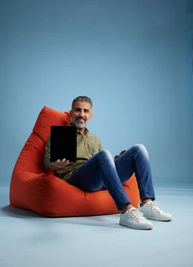 A portrait of a Saudi Arabian Gulf man wearing casual attire, focused while using a tablet while sitting on a chair among orange foliage.