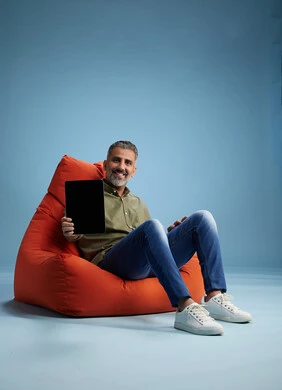 A portrait of a Saudi Arabian Gulf man wearing casual attire, focused while using a tablet while sitting on a chair among orange foliage.