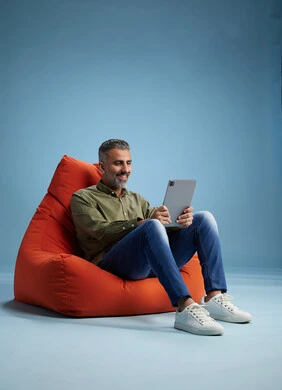 A portrait of a Saudi Arabian Gulf man wearing casual attire, focused while using a tablet while sitting on a chair among orange foliage.