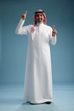 A portrait of a confident Arab Saudi Gulf man wearing a white thobe and a shemagh, smiling, with various gestures and movements, against a blue background.
