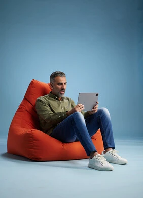 A portrait of a Saudi Arabian Gulf man wearing casual attire, focused while using a tablet while sitting on a chair among orange foliage.