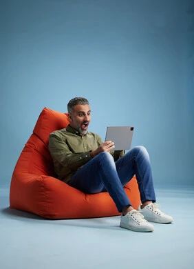 A portrait of a Saudi Arabian Gulf man wearing casual attire, focused while using a tablet while sitting on a chair among orange foliage.