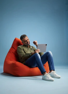 A portrait of a Saudi Arabian Gulf man wearing casual attire, focused while using a tablet while sitting on a chair among orange foliage.