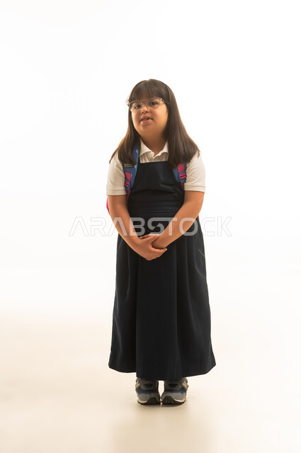 بورتريه خلفية بيضاء , بنت صغيرة عربية خليجية سعودية من ذوي الهمم ترتدي زي مدرسي موحد باللون الرمادي تضع قبضتا اليد امام الجسد , ايماءات السعادة و الابتسامة , الجسم بالطول الكامل