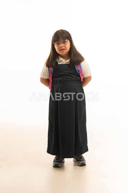 A small Arab Gulf Saudi girl with special needs is standing with her hands clasped behind her back. She is wearing a gray school uniform and carrying a colorful school bag. She has Down syndrome and is depicted in full body against a white background.