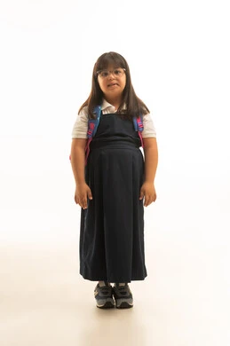 وضع الذراعين بجانب الجسد , بنت صغيرة عربية خليجية سعودية تعاني من متلازمة داون ترتدي الزي المدرسي الموحد لون رمادي تقف امام الكاميرا , حقيبة مدرسية ملونة , الجسم بالطول الكامل , بورتريه خلفية بيضاء