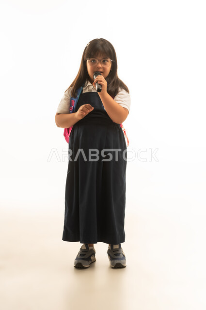 Inclusion of people with determination in creative activities at school, a young Arab Gulf Saudi girl with Down syndrome wearing a gray school uniform holding a megaphone, full-body, portrait, white background, looking at the camera.