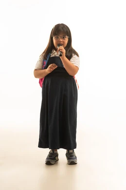 دمج ذوي الهمم في الانشطة الابداعية في المدرسة , بنت صغيرة عربية خليجية سعودية تعاني من متلازمة داون ترتدي الزي المدرسي الموحد باللون الرمادي تحمل مكبر للصوت, الجسم بالطول الكامل , بورتريه خلفية بيضاء , النظر للكاميرا