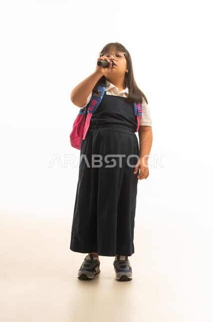 المشاركة في الانشطة الغنائية داخل المدرسة , بنت صغيرة عربية خليجية سعودية تعاني من متلازمة داون ترتدي زي مدرسة موحد باللون الرمادي تحمل مكبر الصوت للغناء , الجسم بالطول الكامل , بورتريه خلفية بيضاء