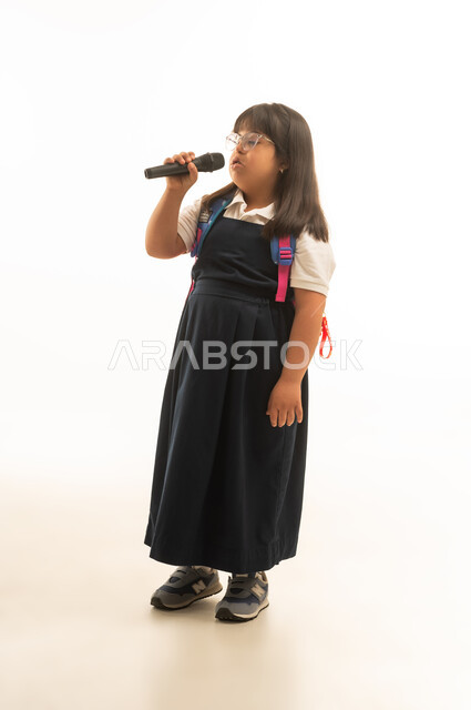 قضاء الاوقات الممتعة و المسلية في الغناء , بنت صغيرة عربية خليجية سعودية تعاني من متلازمة داون ترتدي زي مدرسة موحد لون رمادي تمسك مكبر الصوت , الوقوف بشكل جانبي , الجسم بالطول الكامل , بورتريه خلفية بيضاء