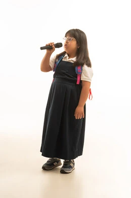 قضاء الاوقات الممتعة و المسلية في الغناء , بنت صغيرة عربية خليجية سعودية تعاني من متلازمة داون ترتدي زي مدرسة موحد لون رمادي تمسك مكبر الصوت , الوقوف بشكل جانبي , الجسم بالطول الكامل , بورتريه خلفية بيضاء