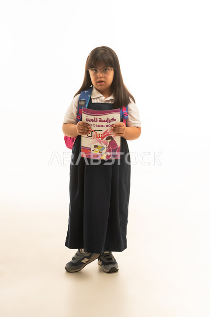 عرض كتاب التلوين امام الكاميرا , بنت عربية خليجية سعودية صغيرة من ذوي الهمم ترتدي الزي المدرسي الموحد باللون الرمادي , الجسم بالطول الكامل , بورتريه خلفية بيضاء
