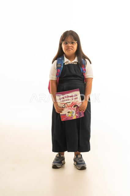 الرسم و التلوين و الانشطة الابداعية لاصحاب متلازمة داون , بنت صغيرة عربية خليجية سعودية من ذوي الهمم ترتدي الزي المدرسي الموحد باللون الرمادي , ايماءات الملل , حمل كتاب التلوين , الجسم بالطول الكامل , بورتريه خلفية بيضاء