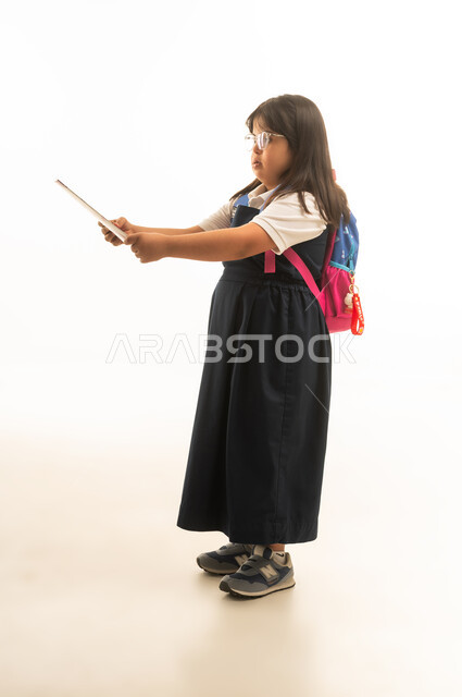 Standing to the side in front of the camera, a young Arab Gulf Saudi girl with Down syndrome wearing a gray school uniform holding study papers, reading a book, full-body shot, portrait with a white background.