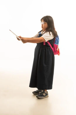 الوقوف بالجانب امام الكاميرا , بنت صغيرة عربية خليجية سعودية تعاني من متلازمة داون ترتدي زي مدرسي موحد باللون الرمادي تحمل الاوراق الدراسية , القراءة في الكتاب , الجسم بالطول الكامل , بورتريه خلفية بيضاء