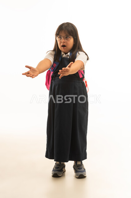 متلازمة داون , بنت صغيرة عربية خليجية سعودية من ذوي الهمم ترتدي الزي المدرسي الموحد باللون الرمادي تفرد الذراعين امام الكاميرا اشارة الى طلب شئ ما , الجسم بالطول الكامل , بورتريه خلفية بيضاء