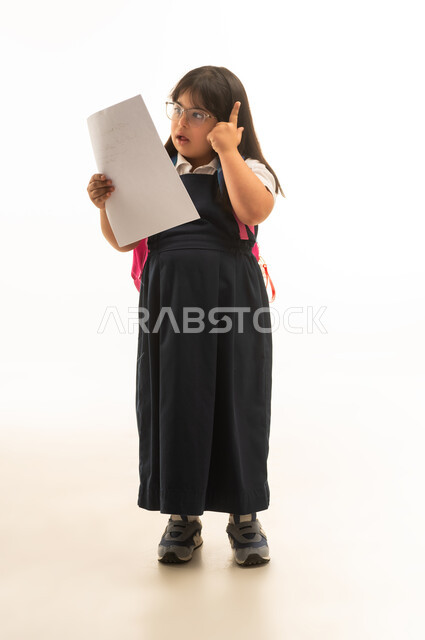 A small Arab Gulf Saudi girl with determination is wearing a gray school uniform, looking at study papers, carrying a colorful school bag, full-length body, portrait with a white background.
