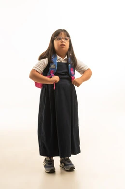 A small Arab Gulf Saudi girl with Down syndrome wearing a gray school uniform, carrying a colorful school bag, pulling the bag's straps, showing signs of boredom, full-body, portrait with a white background.