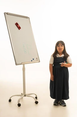 الجسم بالطول الكامل , بورتريه خلفية بيضاء , بنت صغيرة عربية خليجية سعودية تعاني من متلازمة داون ترتدي زي مدرسة موحد باللون الرمادي تمسك القلم الاخضر الخاص بالسبورة البيضاء ,