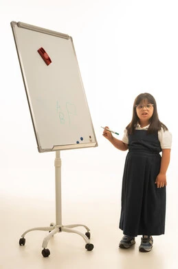 Looking at the camera, a small Arab Gulf Saudi girl with Down syndrome wearing a gray school uniform, standing in front of a whiteboard, full-body shot, portrait with a white background.