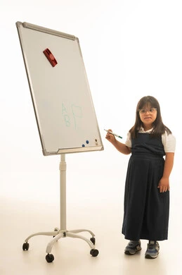 Standing in front of the whiteboard, a small Arab Gulf Saudi girl with Down syndrome is wearing a gray school uniform and holding a colored pencil for the school board. Full-body, portrait with a white background.