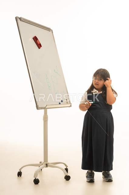 Placing the index finger in the hair is a sign of thinking. A small Arab Gulf Saudi girl with determination wears a gray school uniform and stands in front of a whiteboard, her body in full length, with gestures of questioning and confusion, a portrait with a white background.