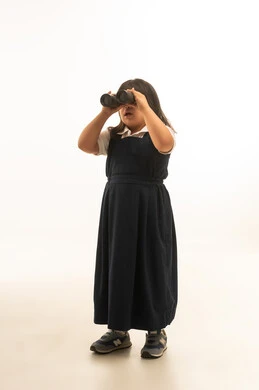 استخدام المنظار المقرب لرؤية شئ ما بعيد , بنت صغيرة عربية خليجية سعودية تعاني من متلازمة داون ترتدي الزي المدرسي الموحد لون رمادي تحمل العدسات المقربة للاشياء ,  الجسم بالطول الكامل , بورتريه خلفية بيضاء