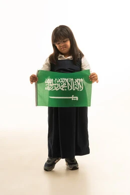 دمج اصحاب متلازمة داون في المجتمع السعودي , بنت صغيرة عربية خليجية سعودية من ذوي الهمم ترتدي زي مدرسة موحد لون رمادي تحمل راية التوحيد الخضراء , النظر الى العلم السعودي بايماءات الحب و الامتنان , الجسم بالطول الكامل , بورتريه خلفية بيضاء