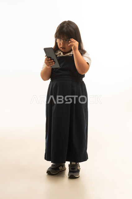 Wearing glasses for clearer vision, a Saudi Arabian Gulf girl with determination wearing a gray school uniform is browsing a mobile phone, full-body shot, portrait with a white background.