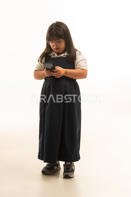 تصفح الهاتف المحمول بسهولة , بنت صغيرة عربية خليجية سعودية من ذوي الهمم ترتدي الزي المدرسي الموحد لون رمادي تستخدم الجهاز التقني, الجسم بالطول الكامل , بورتريه خلفية بيضاء