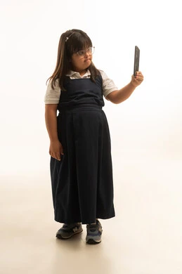 استخدام الهاتف المحمول في التصوير الشخصي , التقاط لقطات مميزة , بنت صغيرة عربية خليجية سعودية تعاني من متلازمة داون ترتدي الزي المدرسي الموحد لون رمادي و نظارات , الجسم بالطول الكامل , بورتريه خلفية بيضاء