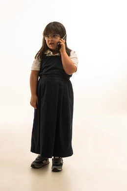 Full body, portrait with a white background, a young Arab Gulf Saudi girl with Down syndrome wearing a gray school uniform, talking on a mobile phone, looking at something.