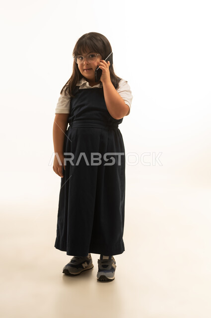 Looking at the camera with gestures of happiness and a smile, a young Arab Gulf Saudi girl with Down syndrome wearing a gray school uniform is talking on a mobile phone, full-body shot, portrait with a white background.