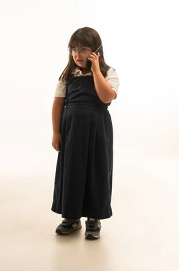 النظر الى شئ ما اثناء التحدث في الهاتف المحمول , بنت صغيرة عربية خليجية سعودية من ذوي الهمم ترتدي زي مدرسي موحد لون رمادي تجري اتصال هاتفي , الجسم بالطول الكامل , بورتريه خلفية بيضاء