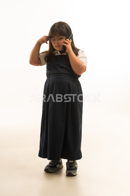 Scratching the head while talking on a mobile phone, a young Saudi Arabian Gulf girl with Down syndrome is talking on the phone, gestures of confusion and questioning, full-body, portrait with a white background.