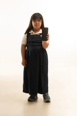 بنت صغيرة عربية خليجية سعوديةتعاني من متلازمة داون ترتدي زي مدرسي موحد لون رمادي تحمل الهاتف المحمول , عرض شاشة سوداء فارغة للكاميرا , الجسم بالطول الكامل , بورتريه خلفية بيضاء