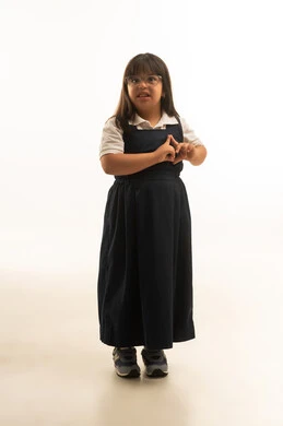 ضم الاصابع على شكل قلب علامة على الحب و الامتنان , بنت صغيرة عربية خليجية سعودية من ذوي الهمم ترتدي زي مدرسة موحد باللون الرمادي تنظر الي الكاميرا , ايماءات السعادة و الابتسامة , الجسم بالطول الكامل , بورتريه خلفية بيضاء