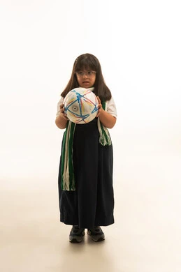 Holding a soccer ball between the hands, looking at something, a small Arab Gulf Saudi girl with Down syndrome wearing a gray school uniform, draping a green scarf over her shoulders, full-body, portrait with a white background.