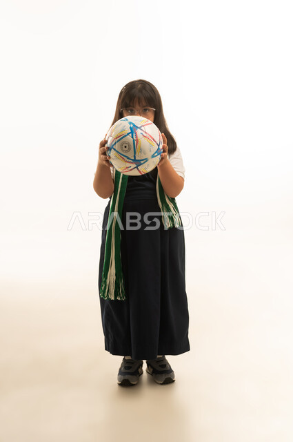 تغطية الوجه بكرة القدم , بنت صغيرة عربية خليجية سعودية من متلازمة داون ترتدي زي مدرسي موحد لون رمادي , وضع كوفية خضراء حول الرقبة , تعابير الحياء و الخجل , الجسم بالطول الكامل , بورتريه خلفية بيضاء