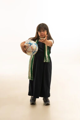 بنت صغيرة عربية خليجية سعودية تعاني من متلازمة داون ترتدي الزي المدرسي الموحد لون رمادي و الكوفية الخضراء تحمل كرة القدم تحت الذراع  , الجسم بالطول الكامل , بورتريه خلفية بيضاء , النظر للكاميرا