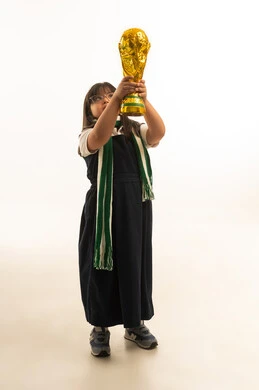 Looking up at something, a small Arab Gulf Saudi girl with Down syndrome wearing a green keffiyeh and a gray school uniform raises the golden cup high after winning in the Paralympic Games, full-body, portrait with a white background.
