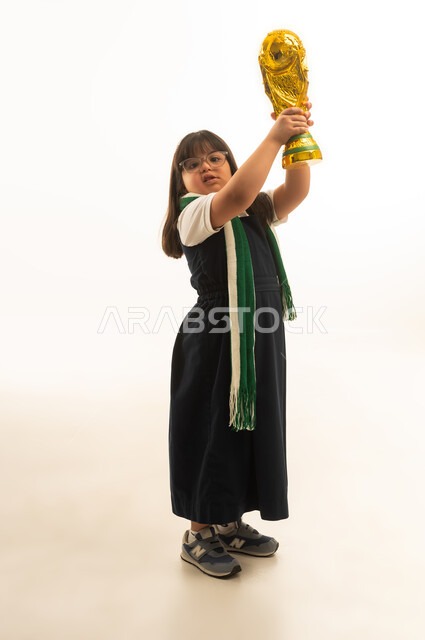 بنت صغيرة عربية خليجية سعودية من ذوي الهمم ترتدي الكوفية الخضراء و الزي المدرسي الموحد لون رمادي ترفع الكأس لاعلى , النظر للكاميرا , بورتريه خلفية بيضاء , الجسم بالطول الكامل